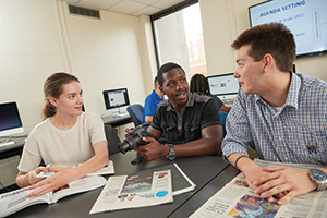 one student is holding camera, newspapers on table, two other students talking