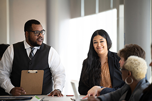 male with clipboard speaks to team sitting at table